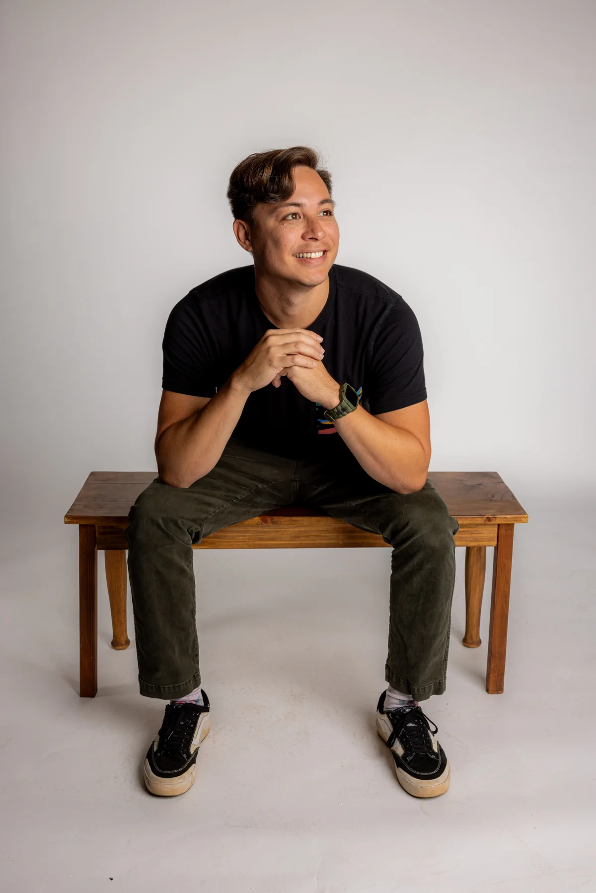 Shane Perry sitting on a wooden bench, smiling in a black T-shirt and olive pants.