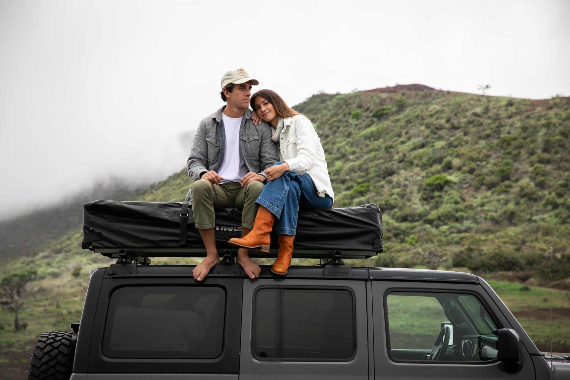 Couple sitting on a Jeep with a rooftop tent, enjoying a scenic mountain view