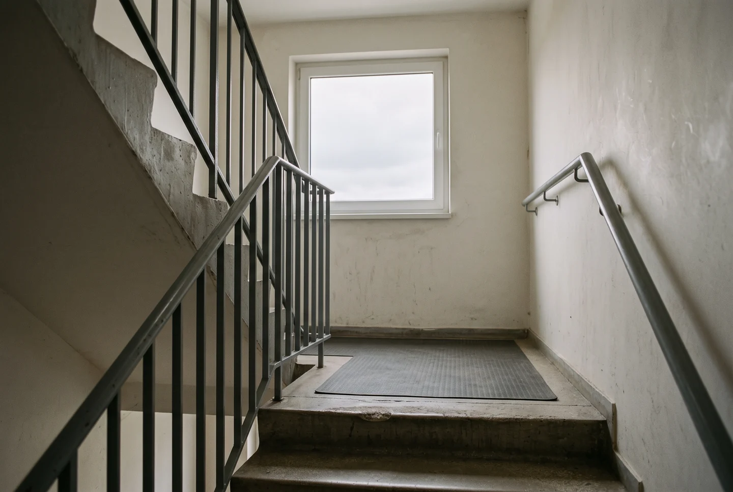 A concrete stairwell half-landing with a plain painted metal handrail and balustrade, a rubber mat on the landing and a single square window onto overcast sky, no people.
