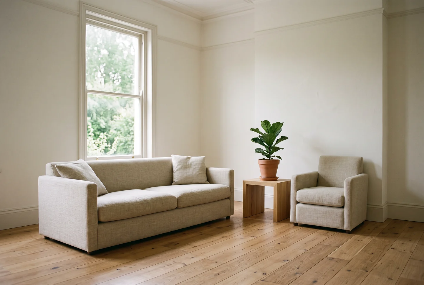 An empty, freshly prepared living room with a pale linen sofa and matching armchair on wide oak floorboards, a small wooden side table with a potted fig, plain white walls and a tall sash window opening onto green trees, no artwork and no people.
