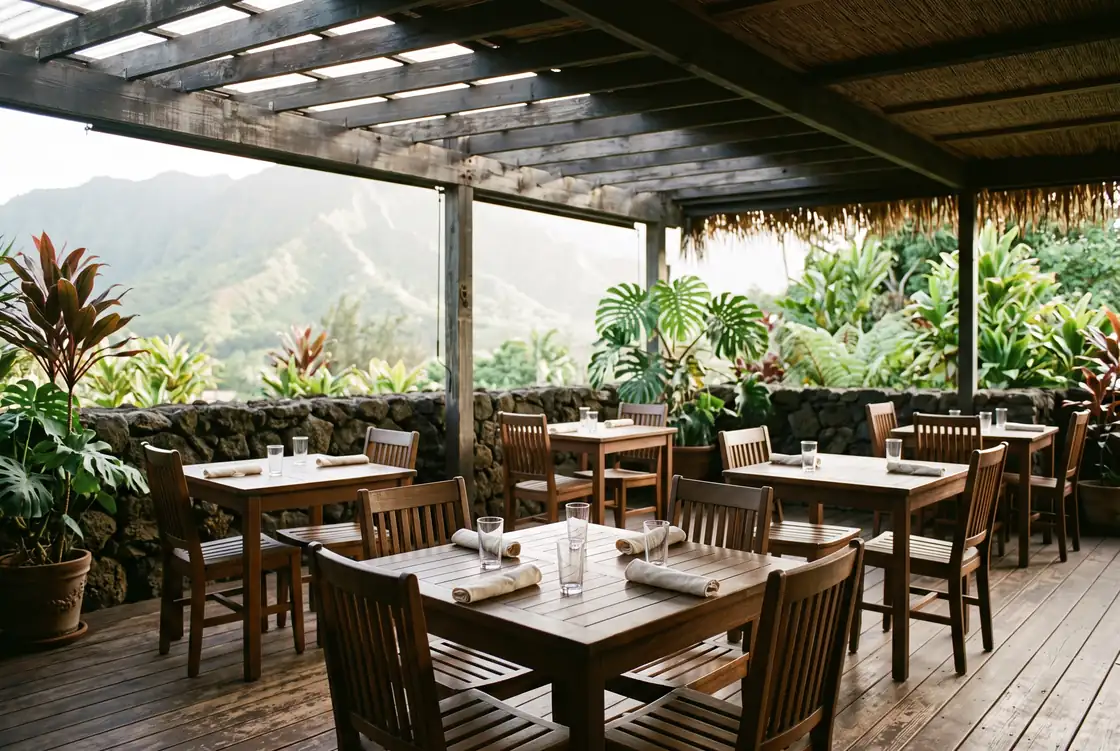 Wide first-light view of an open-air dining terrace with plain wooden tables set with folded cloth napkins and unmarked water glasses, slat-back chairs pushed in, a low lava-rock wall along the open side, potted ti and monstera at the corners, a weathered timber pergola overhead and a green ridge soft in the distance, no people and no printed material on any table.