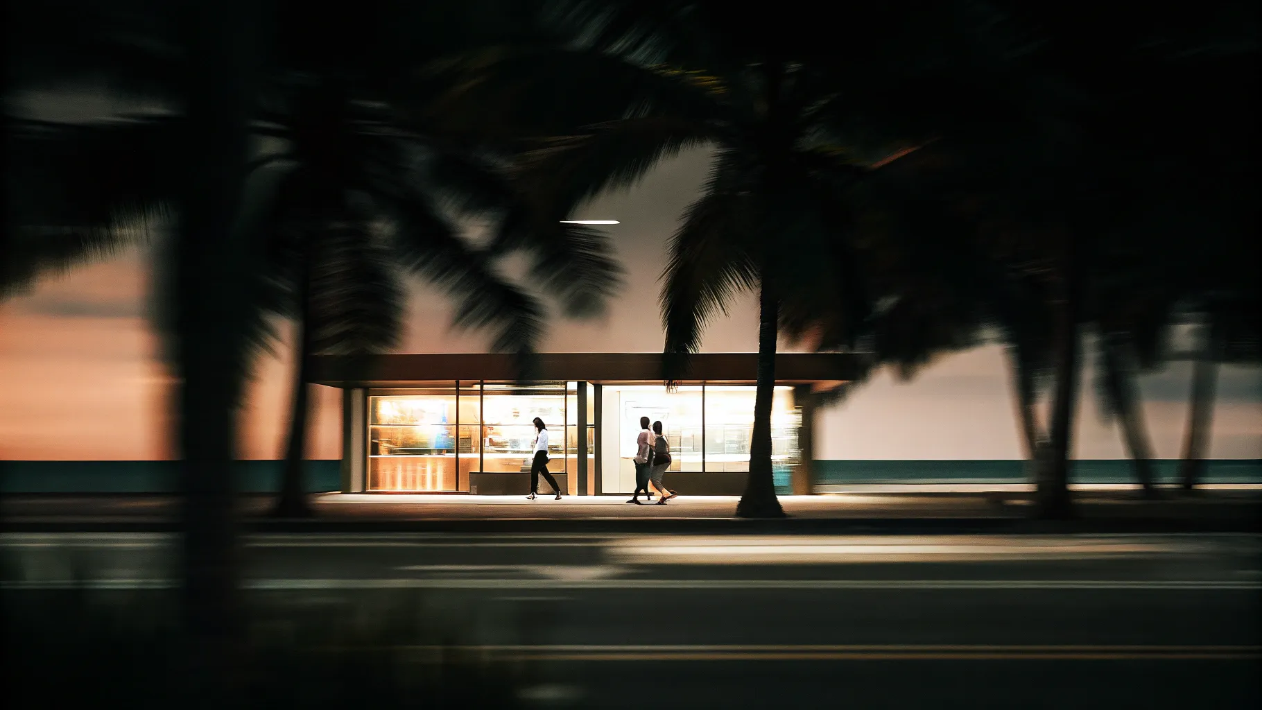 6a4cdf04-4b3b-425c-8162-2a3349945d9c • Digital Reach Digital Reach, Three people stroll past a beachfront kiosk at night, palm trees blurred in the foreground.