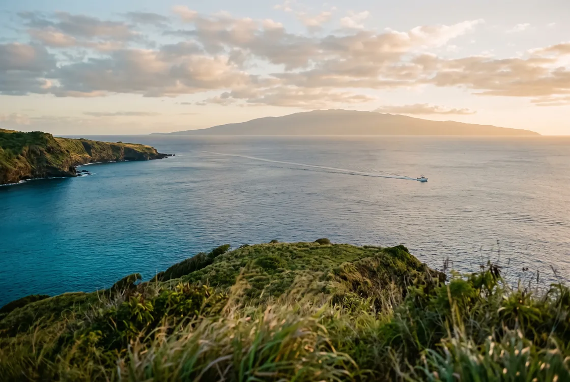 View across a calm inter-island channel from a green headland, with a neighboring Hawaiian island on the horizon and a boat’s wake crossing the water.