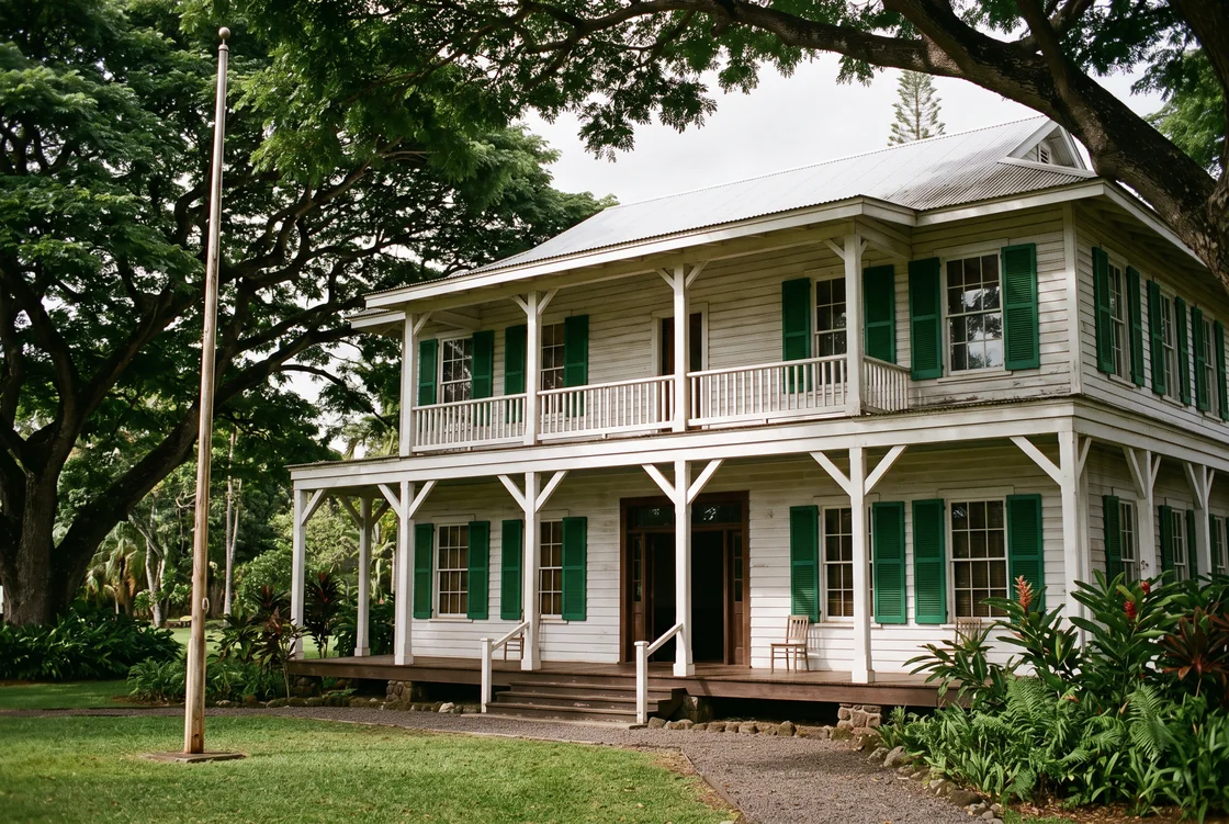 A two-storey plantation-era courthouse with white columns, green shutters and a wide lanai, shaded by large monkeypod trees, an empty flagpole on the lawn.