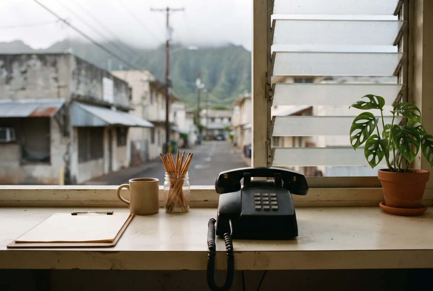 A worn pale laminate front counter in soft morning light with a black corded desk telephone, a blank clipboard, a plain ceramic mug, a jar of pencils and a small potted monstera, a louvered window behind opening onto a narrow street of low buildings with a green mountain ridge beyond, no people.