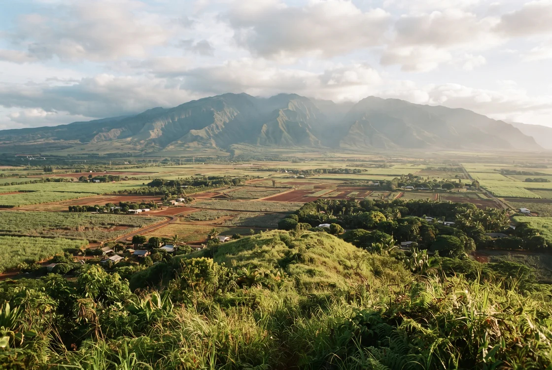 A late-afternoon view from a green foothill ridge across a broad farmed plain toward a second, higher mountain range hazy on the far side, low rooftops half-hidden among trees, red-brown earth between the fields, no people and no vehicles.