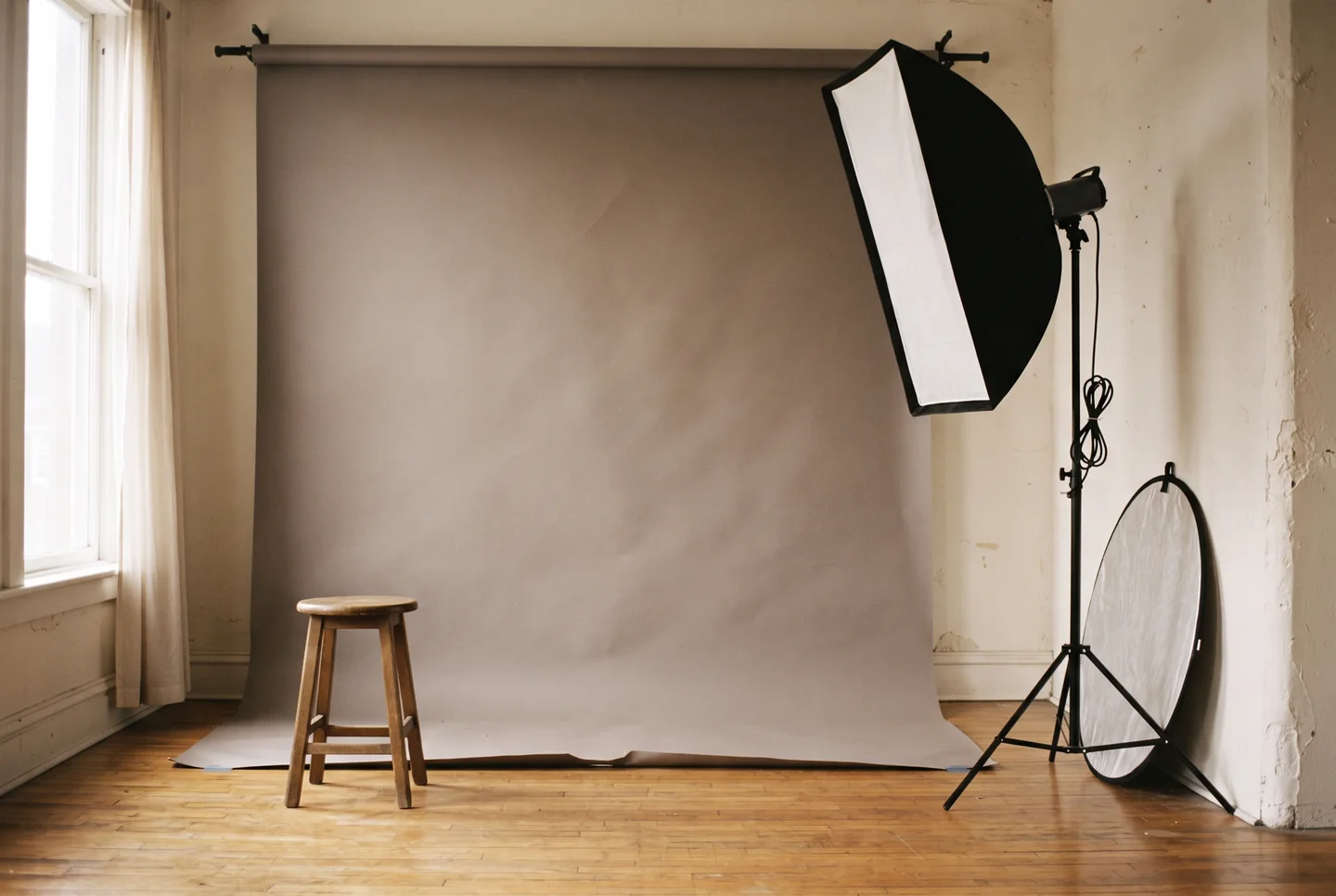 A small portrait studio set for a session: a warm-grey paper backdrop, one softbox on a stand, a wooden stool and a reflector leaning against the wall on a hardwood floor.