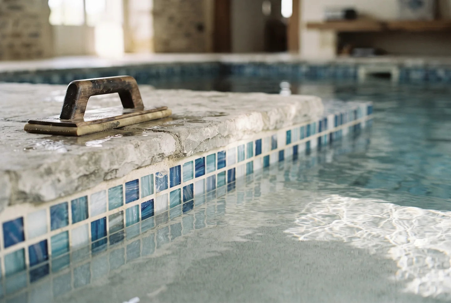 Close detail of a pool edge: glazed blue waterline tile under a stone coping, clear water lapping the tile in low sun.