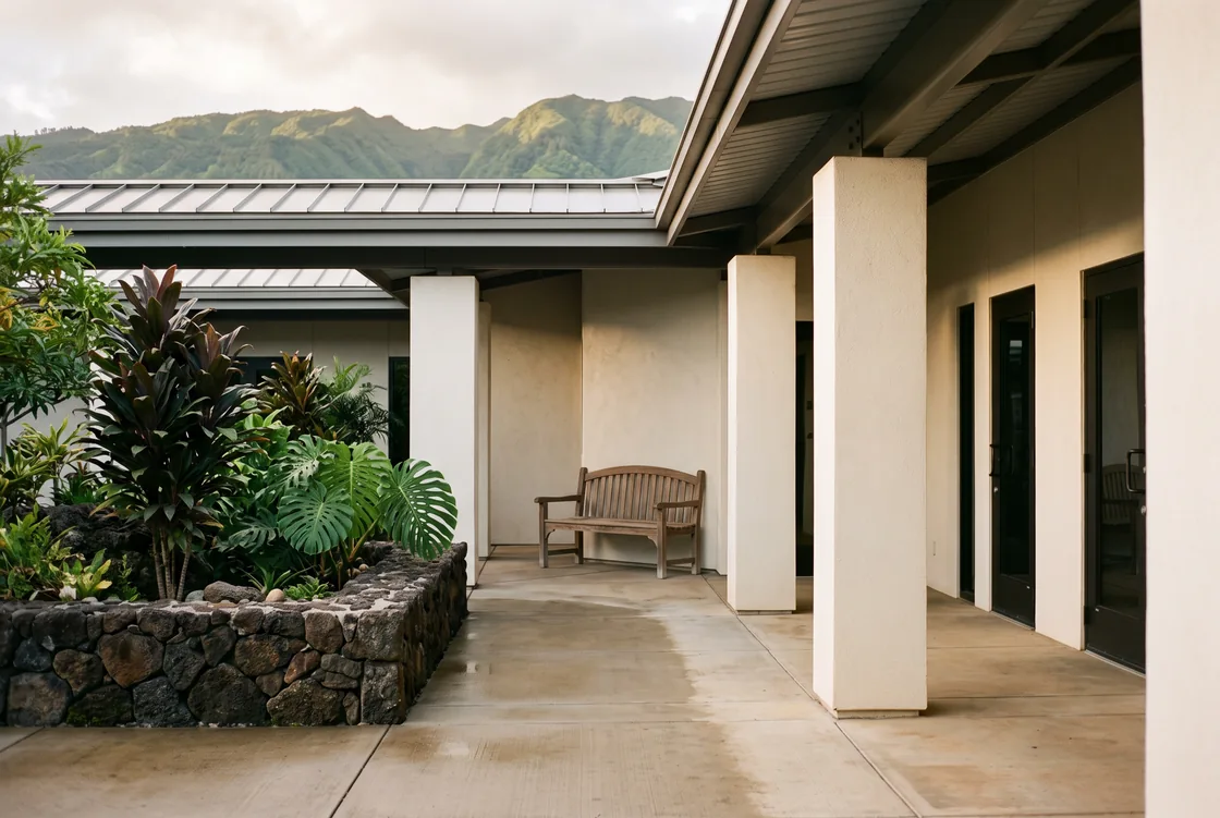 Covered walkway of a low-rise office courtyard in early light: plastered columns, three closed dark-framed glass doors, a wooden bench, a lava-rock planter of ti and monstera, damp pale concrete underfoot and a green ridgeline beyond the metal roof.