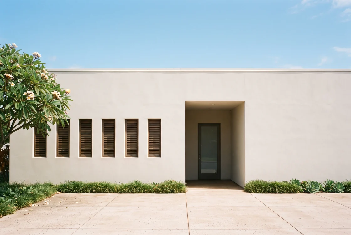 A flat off-white plastered single-storey building front in midday sun with a deep shaded recessed entry and a dark-framed glass door, a run of timber louvred windows, a low strip of clipped green planting, pale concrete paving in front and a flowering plumeria at the left edge, no people and no signage.