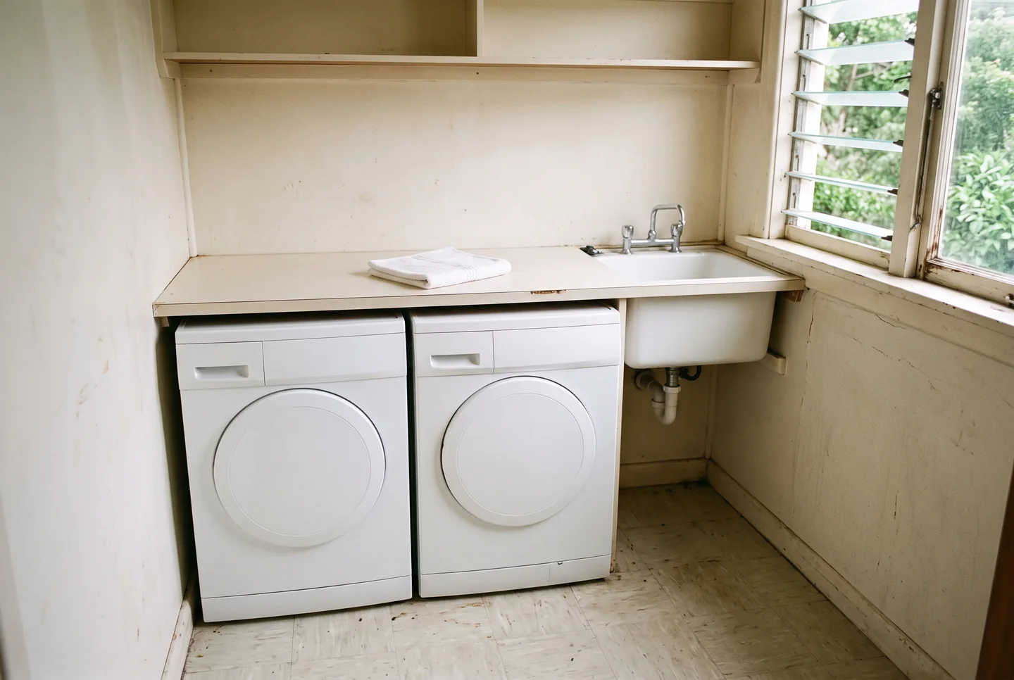 A small laundry alcove with two plain white front-loading machines under a pale laminate counter, a deep white utility sink with a chrome tap, one folded white towel and a louvred window opening onto green foliage, no people.
