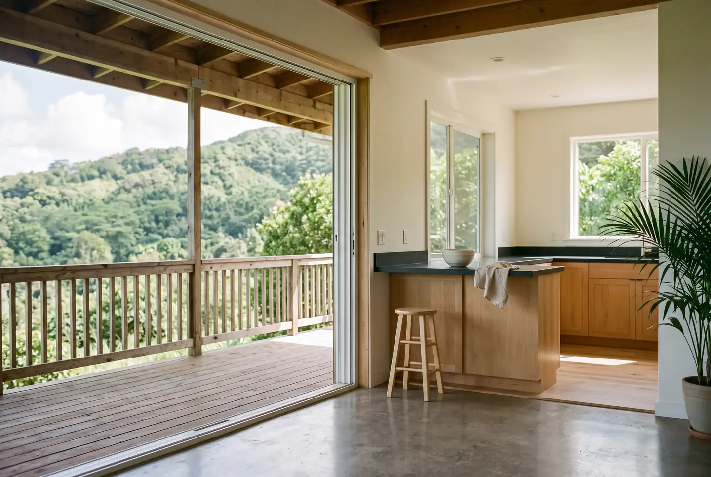 A covered timber lanai with a wooden railing opening through wide sliding doors into a bright empty kitchen with pale wood cabinets, a dark counter with a single bowl and folded cloth, a wooden stool, a potted palm, polished concrete floor and forested hills beyond.